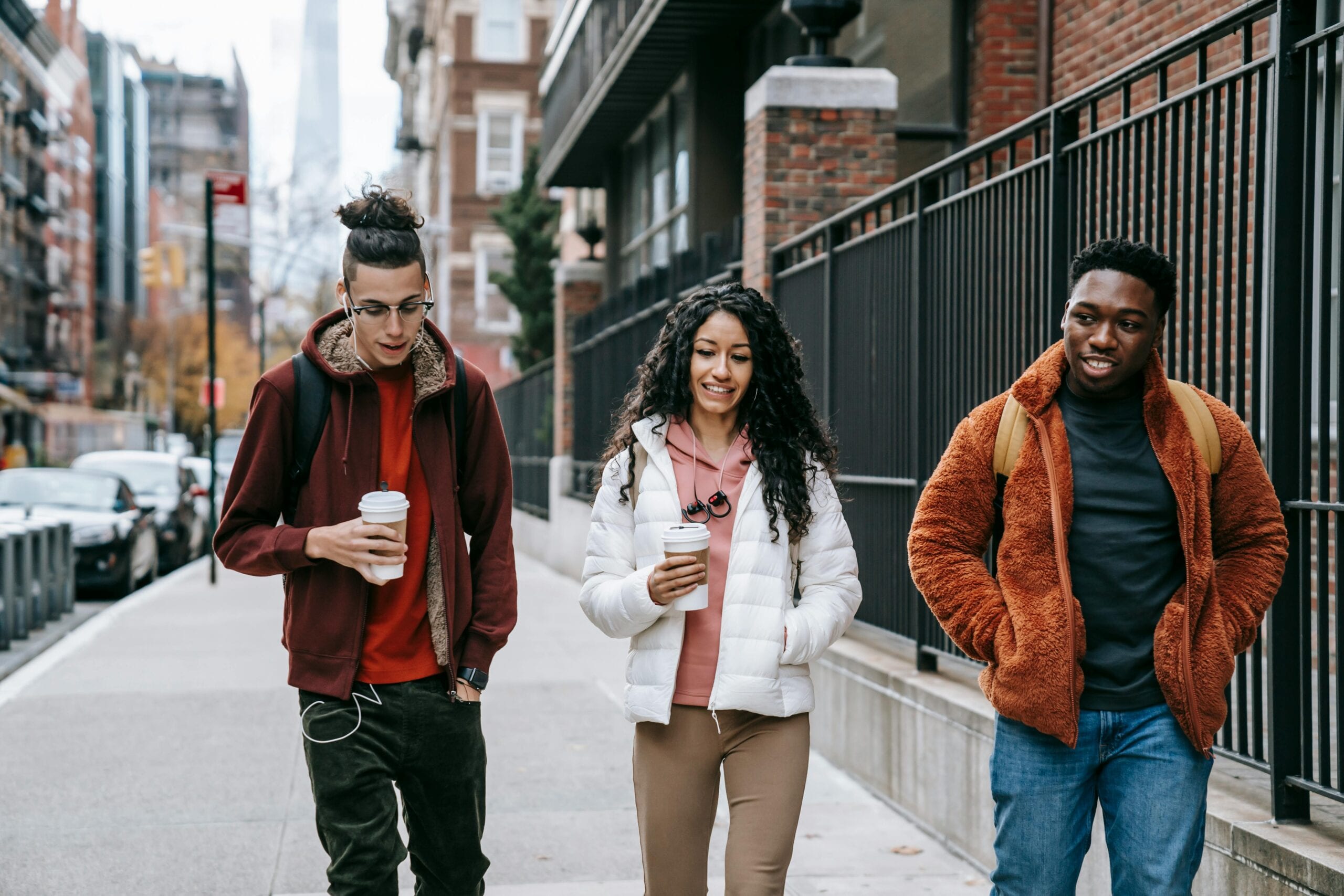 Young glad multiracial partners with hot drinks to go strolling on urban pavement in daytime