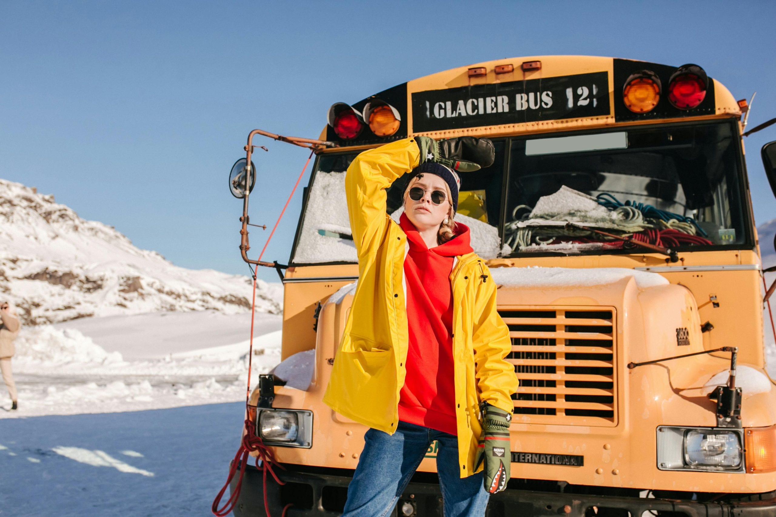 Fashionable woman in yellow coat poses beside glacier bus in snowy winter landscape.