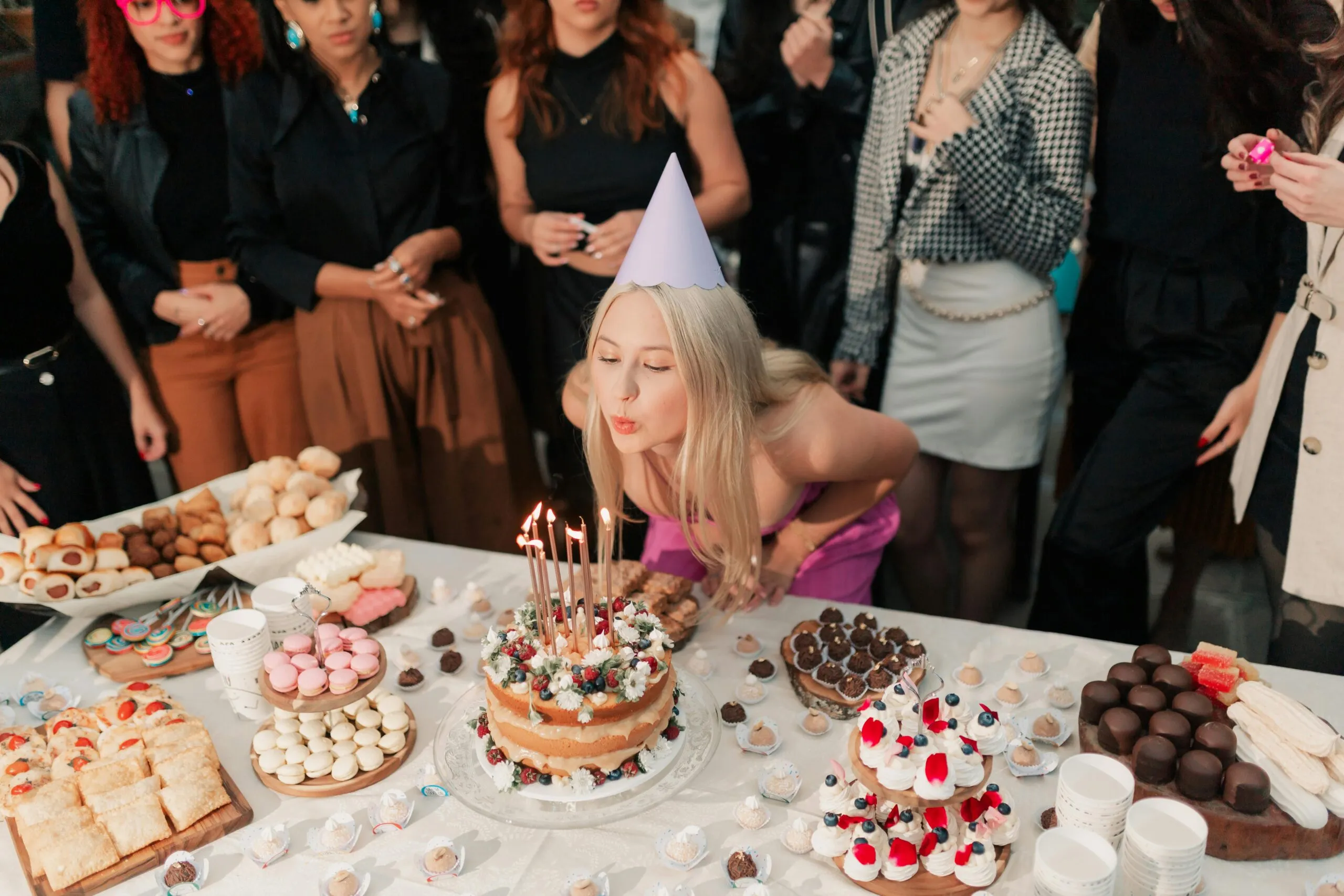 A cheerful young woman celebrates her birthday, surrounded by friends and a variety of delicious cakes and desserts.